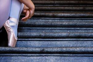 Nahansicht von Ballerina tragen Punkte Schuhe halten ihr Hände auf ihr Knöchel während Sitzung auf das grau Stein Schritte. tanzen ruhen auf das Treppe. Tänzer Füße mit Rosa Ballett Hausschuhe An. Kopieren Raum foto