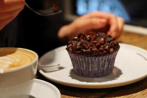 Hand von jung Frau halten Gabel und Essen Cupcake. Tasse von Kaffee Latté auf das Tisch. hausgemacht Schokolade Muffin oder Brownie auf ein Weiß Platte. Frau Frau Trinken Cappuccino. selektiv Fokus. foto