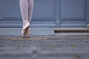 Nahansicht von ein Tänzer Beine balancieren auf das pointe Schuhe. Ballerina tragen Ballett Hausschuhe während Stehen auf ihr Tipps auf das Kopfsteinpflaster Straße. grau retro Türen auf das Hintergrund. selektiv Fokus foto