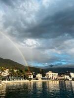 Regenbogen Über das Häuser auf das Meer Küste beim das Fuß von das Berge gegen ein wolkig Himmel foto