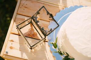 Paar von Engagement Ringe Lügen im ein Glas Box Nächster zu ein Hochzeit Kuchen auf ein hölzern Box foto