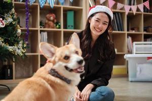 Eine junge asiatische schöne junge Arbeiterin in einem roten Hut neckt einen Hund mit Liebe auf einer Geschäftsbüroparty, dekorativ für das Feiern des Weihnachtsfestes und der Neujahrsfeiertage. foto