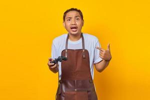 Porträt eines überraschten hübschen asiatischen jungen Mannes mit Schürze, der einen Gamecontroller hält und auf sich selbst über einem gelben Hintergrund zeigt foto