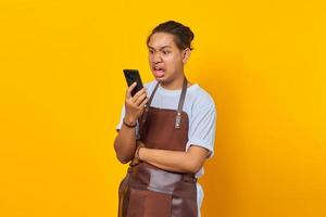 Porträt eines jungen asiatischen Mannes mit Schürze, der wütend aussieht, während er auf dem Smartphone auf gelbem Hintergrund spricht foto