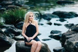 blondes Mädchen in einem schwarzen Kleid mit blauen Augen an der Küste des Flusses foto
