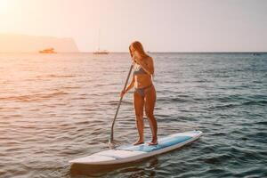 Frau Meer sup. schließen oben Porträt von glücklich jung kaukasisch Frau mit lange Haar suchen beim Kamera und lächelnd. süß Frau Porträt im ein Blau Bikini posieren auf sup Tafel im das Meer foto