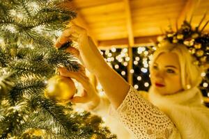 ein blond Frau im Weiß Kleid und ein Krone von Gold Ornamente schmücken Weihnachten Baum mit Gold Ornamente und Beleuchtung. das Baum ist dekoriert mit Gold Bälle und ist zündete oben mit Beleuchtung. foto