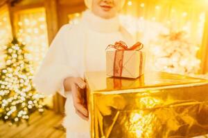 Frau im Weiß Mantel halten goldverpackt Geschenk Box mit rot Band im Vorderseite von Weihnachten Baum, implizieren Urlaub Jahreszeit Geschenk Austausch. foto