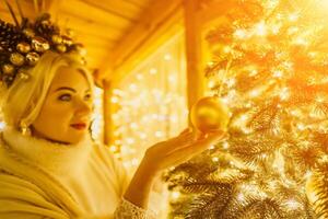 ein blond Frau im Weiß Kleid und ein Krone von Gold Ornamente schmücken Weihnachten Baum mit Gold Ornamente und Beleuchtung. das Baum ist dekoriert mit Gold Bälle und ist zündete oben mit Beleuchtung. foto