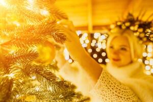 ein blond Frau im Weiß Kleid und ein Krone von Gold Ornamente schmücken Weihnachten Baum mit Gold Ornamente und Beleuchtung. das Baum ist dekoriert mit Gold Bälle und ist zündete oben mit Beleuchtung. foto