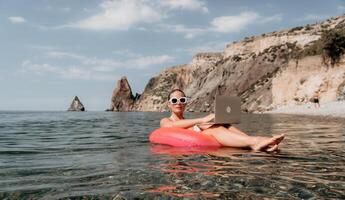 Frau Freiberufler funktioniert auf Laptop Schwimmen im Meer auf Rosa aufblasbar Ring. glücklich Tourist im Sonnenbrille schwebend auf aufblasbar Krapfen und Arbeiten auf Laptop Computer im Ruhe Ozean. Fernbedienung Arbeiten irgendwo foto