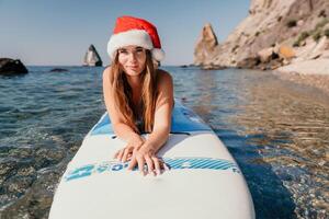 Frau Meer sup. schließen oben Porträt von glücklich jung kaukasisch Frau mit lange Haar im Santa Hut suchen beim Kamera und lächelnd. süß Frau Porträt im ein Weiß Bikini posieren auf sup Tafel im das Meer foto