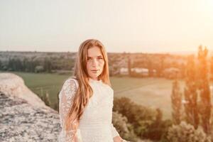 glücklich Frau im Weiß Boho Kleid auf Sonnenuntergang im Berge. romantisch Frau mit lange Haar Stehen mit ihr zurück auf das Sonnenuntergang im Natur im Sommer- mit öffnen Hände. Silhouette. Natur. Sonnenuntergang. foto