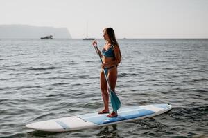 Frau Meer sup. schließen oben Porträt von glücklich jung kaukasisch Frau mit lange Haar suchen beim Kamera und lächelnd. süß Frau Porträt im ein Blau Bikini posieren auf sup Tafel im das Meer foto