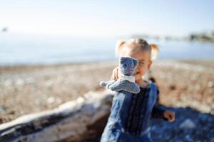 wenig lächelnd Mädchen steht in der Nähe von ein Treibholz auf ein sonnig Strand und hält aus ein Spielzeug foto