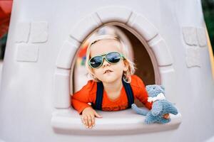 wenig Mädchen im Sonnenbrille mit ein Spielzeug im ihr Hand sieht aus aus von das Fenster von ein Spielzeug Schloss auf das Spielplatz foto