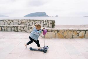 wenig Mädchen Fahrten ein Roller auf ein gepflastert Bereich in der Nähe von das Meer suchen Nieder beim ihr Füße foto