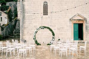 runden Hochzeit Bogen steht im Vorderseite von Reihen von Weiß Stühle in der Nähe von ein Kirche foto