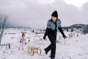 Mama ist ziehen ein Schlitten auf ein Seil oben das Berg, suchen zurück beim das wenig Mädchen folgenden hinter ihr foto