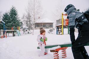 Mama und wenig Mädchen schwingen auf ein schneebedeckt Schaukel-Balancer beim das Spielplatz foto