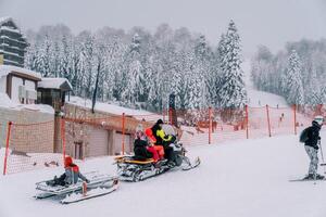Papa und Kinder Reiten ein Schneemobil mit ein Schlitten befestigt auf ein schneebedeckt Berg entlang ein rot Zaun foto