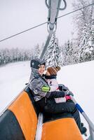 lächelnd Mutter mit ein wenig Mädchen im ihr Waffen Fahrten ein Sessellift oben das Berg foto