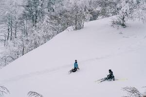 Snowboarder kniet in der Nähe von ein Skifahrer Sitzung im das Schnee auf ein Berg Steigung foto