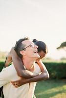 Lachen Mann trägt Frau auf seine zurück über Grün Rasen foto