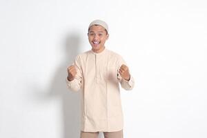 Porträt von religiös asiatisch Muslim Mann im koko Hemd mit Schädeldecke erziehen seine Faust, feiern Erfolg. Leistung Konzept. isoliert Bild auf Weiß Hintergrund foto