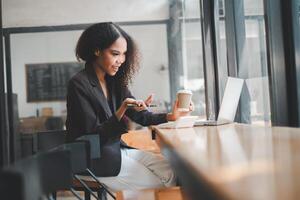 ein aufmerksam Geschäftsfrau mit ein Kaffee Tasse im einer Hand und ein Stift im das andere funktioniert fleißig auf ihr Laptop im das beschwingt Atmosphäre von ein Cafe foto