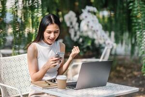 ein jung Frau reagiert mit Freude zu ihr Smartphone während nehmen ein brechen von ihr Laptop Arbeit beim ein Garten Tisch, ein Kaffee Tasse innerhalb einfach erreichen. foto