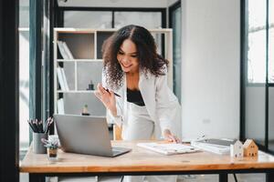 ein charismatisch Geschäftsfrau Gesten während ein animiert Gespräch, wahrscheinlich auf ein Video Forderung, wie sie steht beim ihr Arbeit Schreibtisch im ein hell, zeitgenössisch Büro Raum. foto