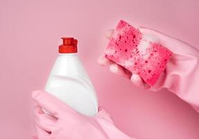 ein Frau Hände im ein Rosa Gummi Handschuhe halt ein Seife Küche Schwamm und Waschmittel auf ein Rosa Hintergrund. Geschirrspülen Konzept. Nahansicht. selektiv Fokus. foto