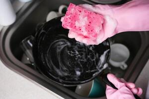 ein Frau im Gummi Handschuhe wäscht ein braten schwenken im das Küche mit Waschmittel. Nahansicht. selektiv Fokus. foto