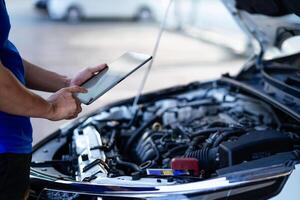 ein Mann im ein Blau Hemd ist suchen beim ein Tablette während Stehen Nächster zu ein Wagen. er ist Überprüfung das Autos Motor foto