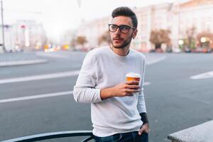 Hipster Mann Stehen mit wegbringen Kaffee, lächelnd angenehm, Gehen auf th Stadt Straße. glücklich sorglos gut aussehend Kerl im Brillen foto