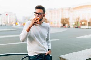 Hipster Mann Stehen mit wegbringen Kaffee, lächelnd angenehm, Gehen auf th Stadt Straße. glücklich sorglos gut aussehend Kerl im Brillen foto