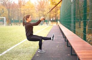 Mädchen Athlet funktional Ausbildung auf Sportplatz. gemischt Rennen jung Erwachsene Frau tun trainieren mit Suspension System. gesund Lebensstil. Dehnen draußen Spielplatz. foto