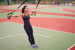 Mädchen Athlet Ausbildung mit trx auf Sportplatz. gemischt Rennen jung Erwachsene Frau tun trainieren mit Suspension System. gesund Lebensstil. Dehnen draußen Spielplatz. foto