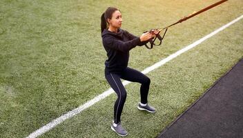 Mädchen Athlet Ausbildung mit trx auf Sportplatz. gemischt Rennen jung Erwachsene Frau tun trainieren mit Suspension System. gesund Lebensstil. Dehnen draußen Spielplatz. foto