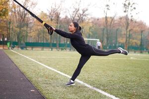 Mädchen Athlet Ausbildung mit trx auf Sportplatz. gemischt Rennen jung Erwachsene Frau tun trainieren mit Suspension System. gesund Lebensstil. Dehnen draußen Spielplatz. foto