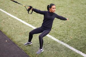 Mädchen Athlet Ausbildung mit trx auf Sportplatz. gemischt Rennen jung Erwachsene Frau tun trainieren mit Suspension System. gesund Lebensstil. Dehnen draußen Spielplatz. foto