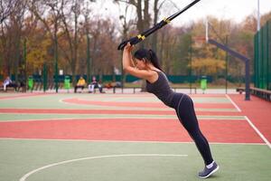 Mädchen Athlet funktional Ausbildung auf Sportplatz. gemischt Rennen jung Erwachsene Frau tun trainieren mit Suspension System. gesund Lebensstil. Dehnen draußen Spielplatz. foto