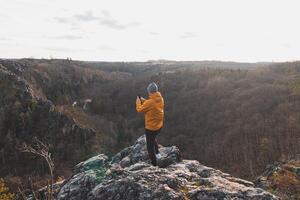 Reisender im ein Gelb Jacke Stehen auf das Kante von ein Cliff nehmen ein Bild von das Landschaft auf seine Handy, Mobiltelefon Telefon beim Sonnenuntergang. ein Erinnerung von ein Reise. wild sarka Schlucht, Prag foto