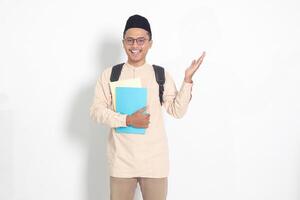 Porträt von aufgeregt Schüler asiatisch Muslim Mann im koko Hemd mit Schädeldecke Tragen Rucksack, halten Schule Bücher, zeigen zu das Seite. islamisch Bildung Konzept. isoliert Bild auf Weiß Hintergrund foto