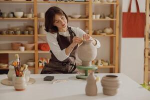 lächelnd jung weiblich Keramiker tragen Schürze Arbeit mit ungebrannt Lehm Vase im Keramik Studio foto