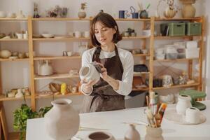 lächelnd jung weiblich Keramiker tragen Schürze Arbeit mit ungebrannt Lehm Vase im Keramik Studio foto