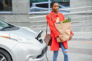 Laden Elektro Auto beim das elektrisch Gas Bahnhof. afrikanisch amerikanisch Mädchen Stehen durch das Auto foto