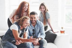 Kerl mit langen Haaren, der lustiges Material auf seinem Handy zeigt. Bürofreunde haben fröhliche Zeit auf dem weißen Sofa zu sitzen foto