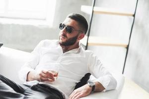 nachdenklicher Blick. Foto eines jungen bärtigen Geschäftsmannes mit Sonnenbrille und Whisky in der Hand sitzen auf dem weißen Sofa